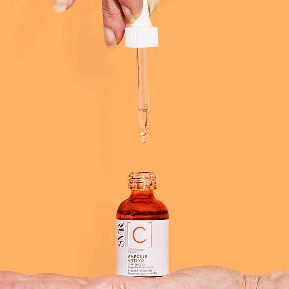 Hand holding an amber glass bottle of SVR Ampoule Anti-Ox serum with a white dropper dispensing clear liquid on orange.