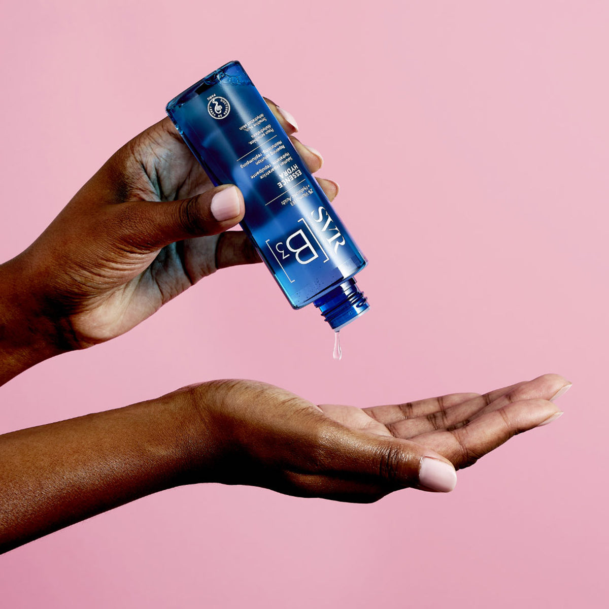 Person pouring blue SVR B3 Hydra Essence from a clear plastic bottle into their palm against a pink background.