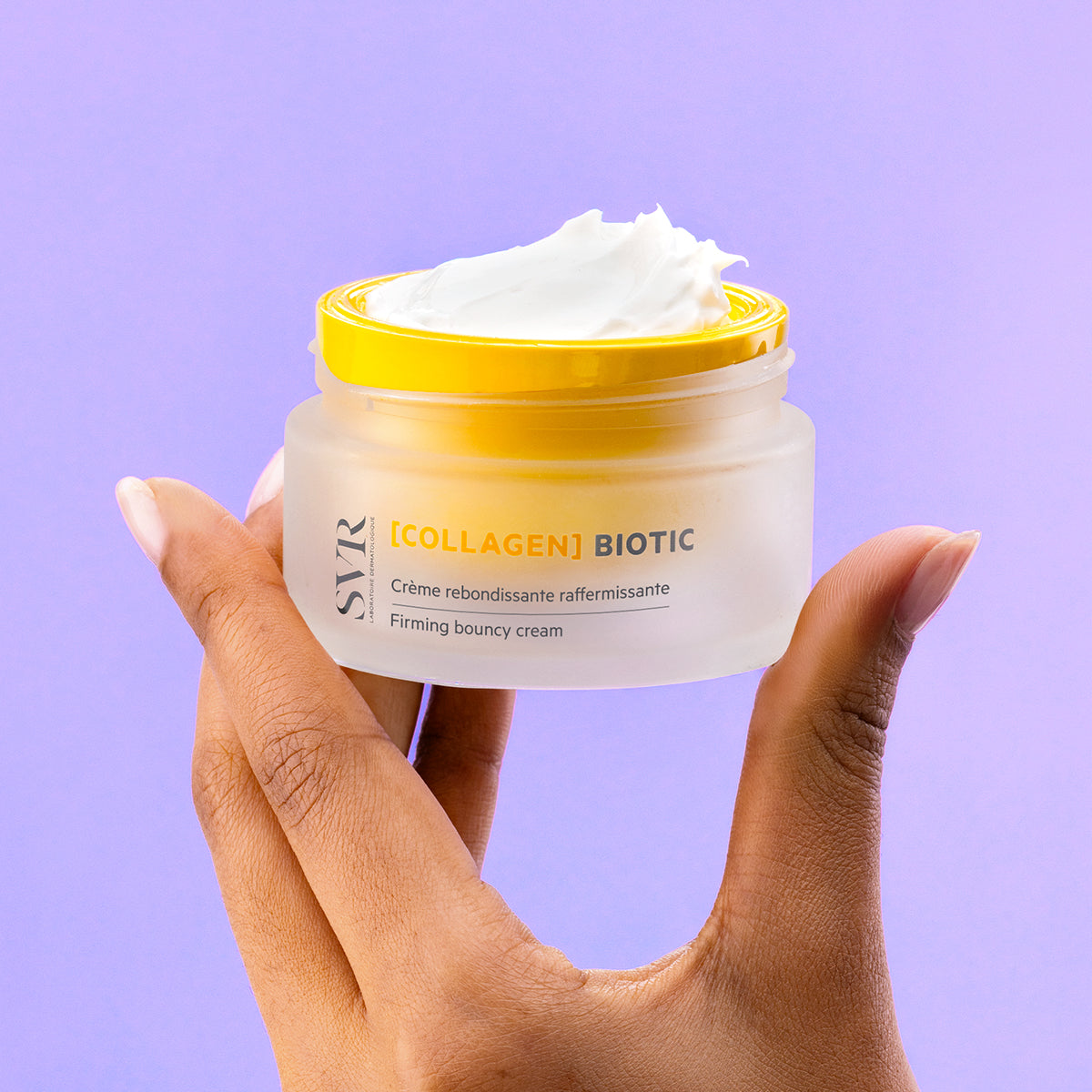 Hand holding a frosted glass jar of SVR Collagen Biotic firming cream with a bright yellow lid against a purple background.