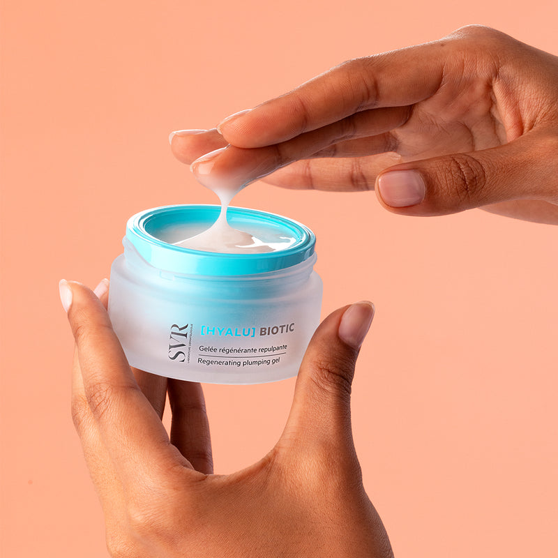 Hands holding a frosted glass jar of SVR Hyalu Biotic plumping gel with a bright blue lid against a peach background.