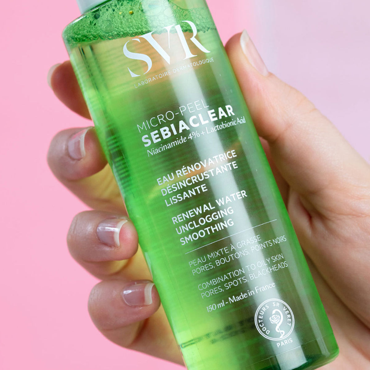Hand holding a transparent green 150ml bottle of SVR Sebiaclear Micro-Peel Essence against a pink background.