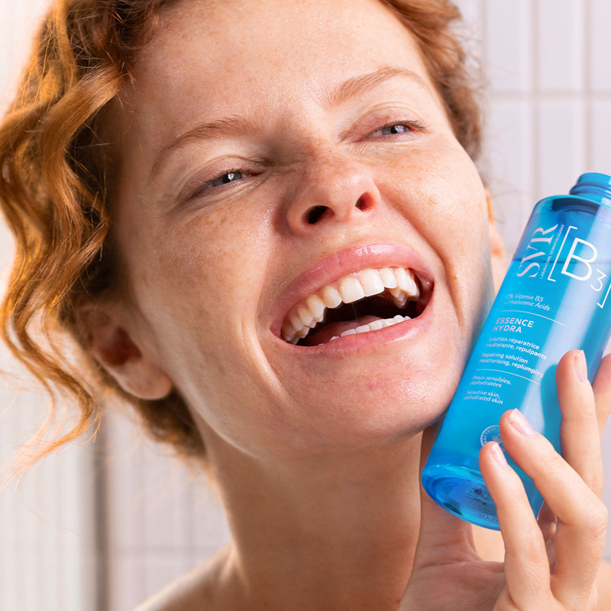 Smiling woman holding a blue translucent bottle of SVR B3 Hydra Essence skincare for dehydrated skin.