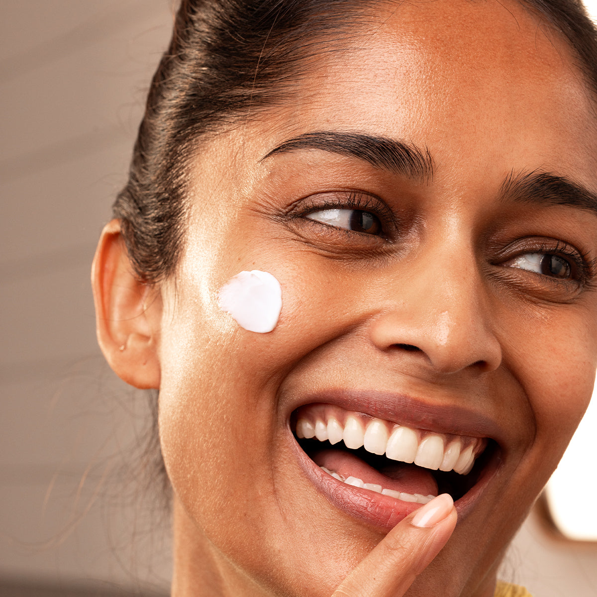 Smiling woman with radiant skin applying a dab of white Collagen Biotic cream to her cheek for a firm, hydrated look.