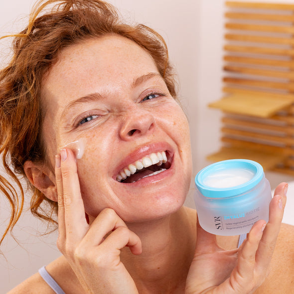 Smiling woman applying SVR HYALU Biotic regenerating plumping gel from a frosted glass jar with a light blue lid.
