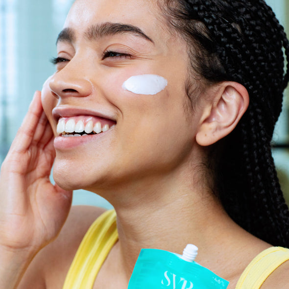 Smiling woman with braids applying white moisturizing cream from a teal SVR pouch to her cheek for hydrated skin.
