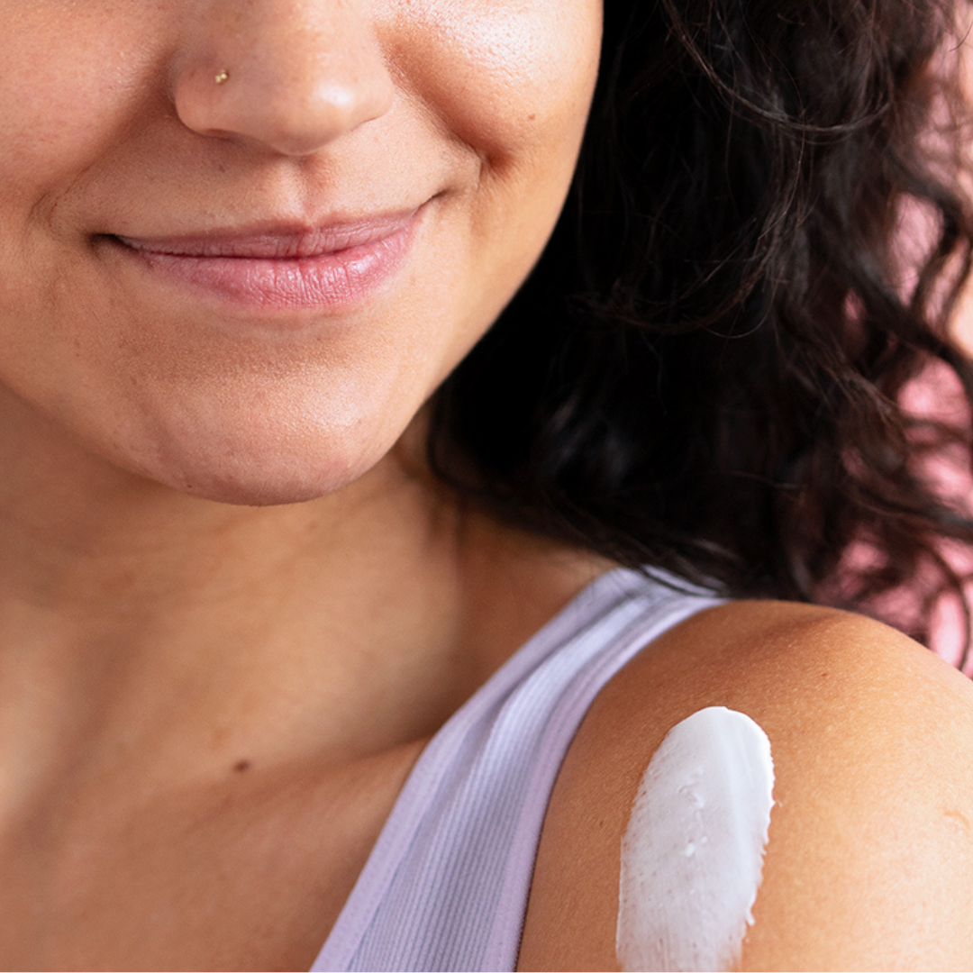 Smiling woman with curly hair applying a smooth, white TOPIALYSE nourishing cream to her shoulder against a pink background.