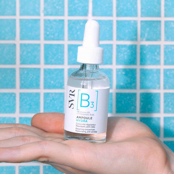 SVR Ampoule B3 Hydra serum bottle with a white dropper cap held in a hand against a blue tiled background.