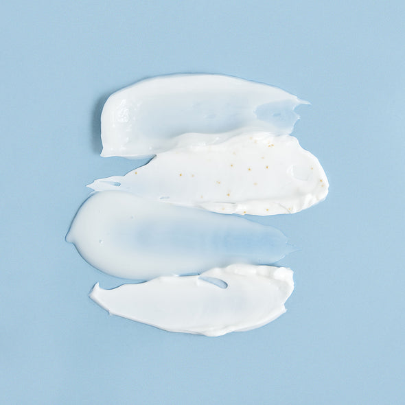 Three smooth, white cream and gel swatches with tiny gold particles layered on a soft blue background.