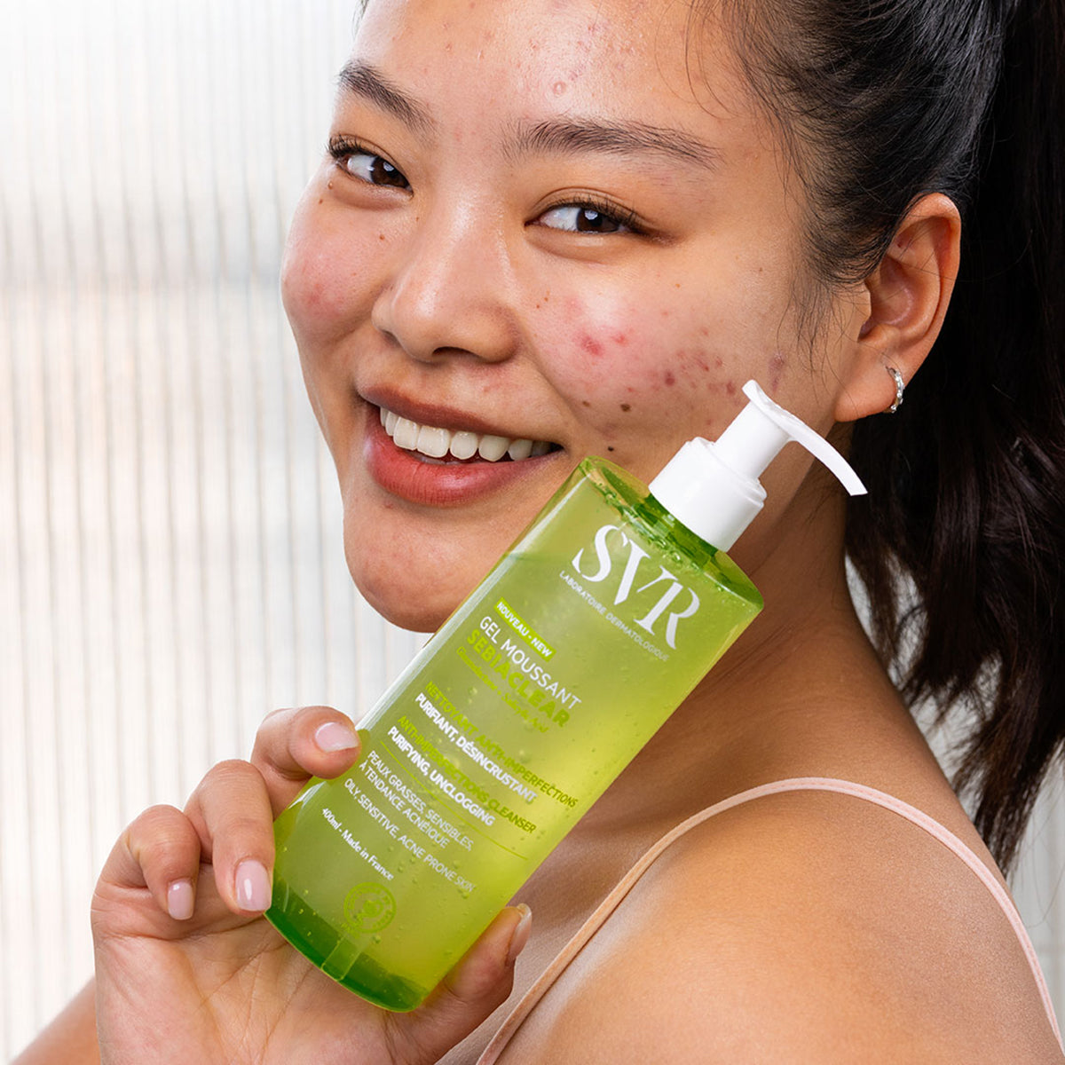 Smiling woman holding a 400ml clear green SVR Sebiaclear foaming gel pump bottle for acne-prone skin.