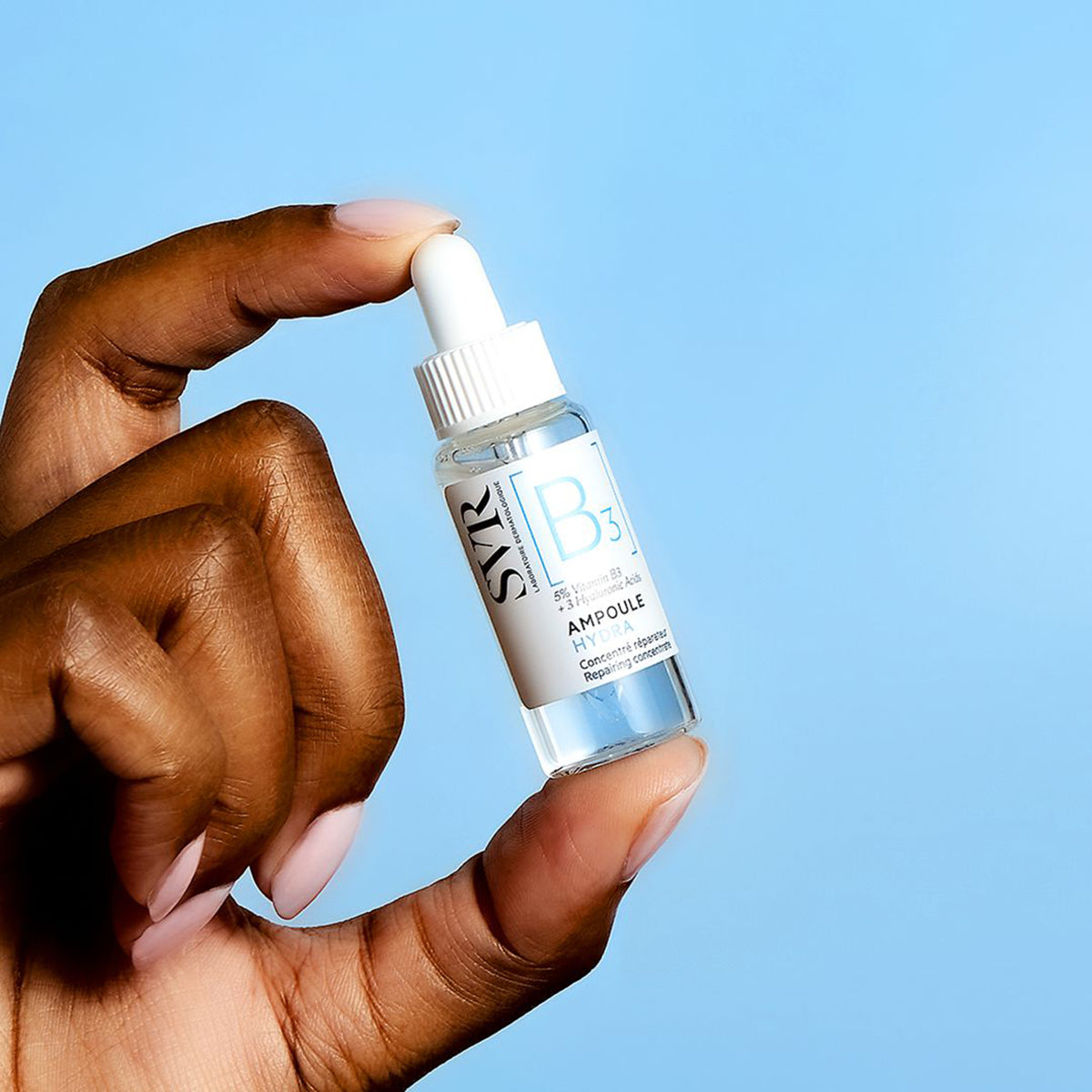 Hand holding a clear glass SVR Ampoule B3 Hydra serum bottle with a white dropper cap against a light blue background.