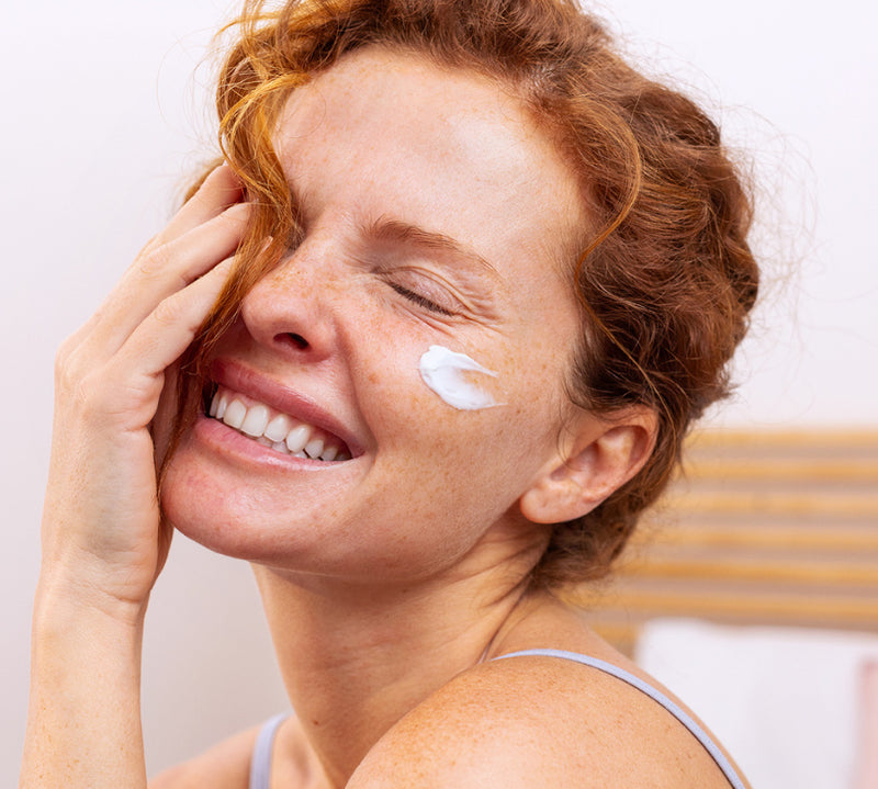 Red-haired woman smiling with white collagen cream applied on her cheek in a bright room.