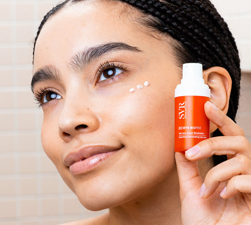 Woman applying white cream under eye holding orange SVR Vitamin C Eye Biotic bottle.