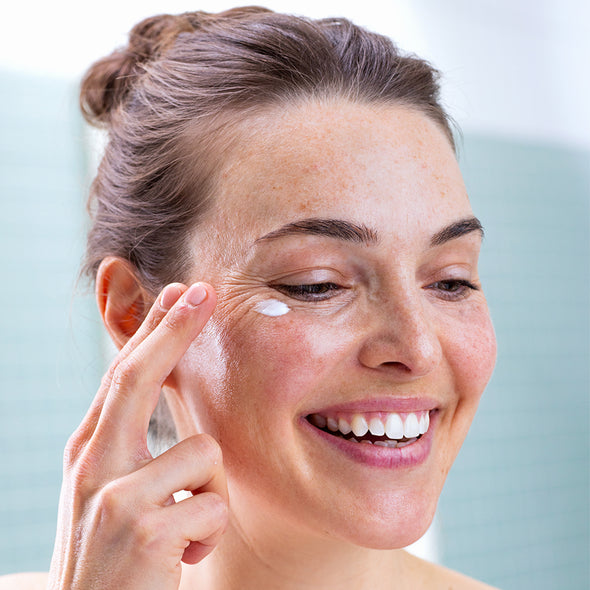 Smiling woman applying white SVR Palpebral eye cream to her sensitive under-eye area with her fingertip.