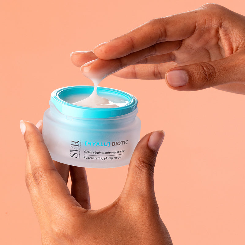Hands holding a frosted glass jar of SVR HYALU Biotic gel with a bright blue lid and a dollop of white cream.