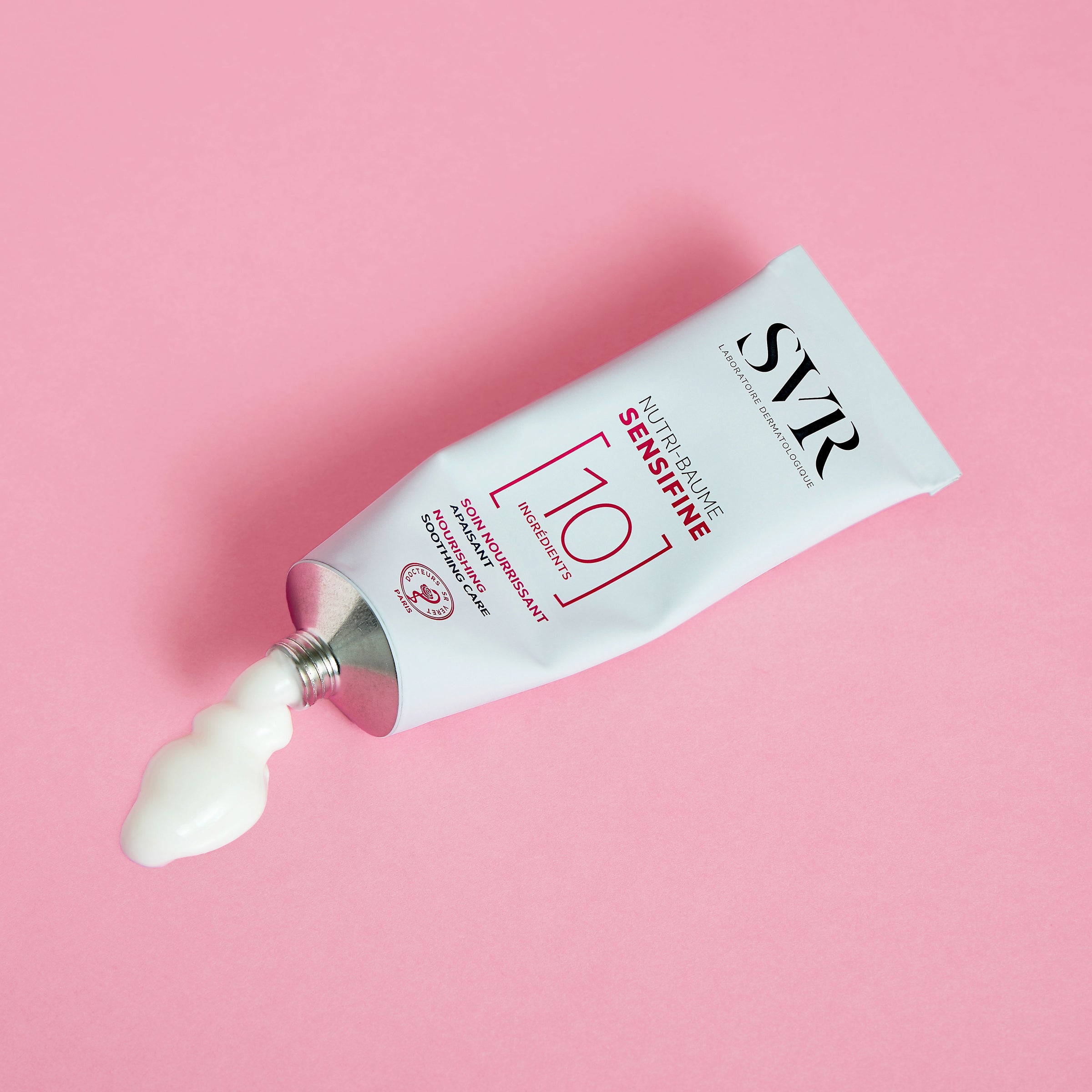SVR Sensifine Nutri-Baume white aluminum tube squeezing out rich cream on a solid pink background.