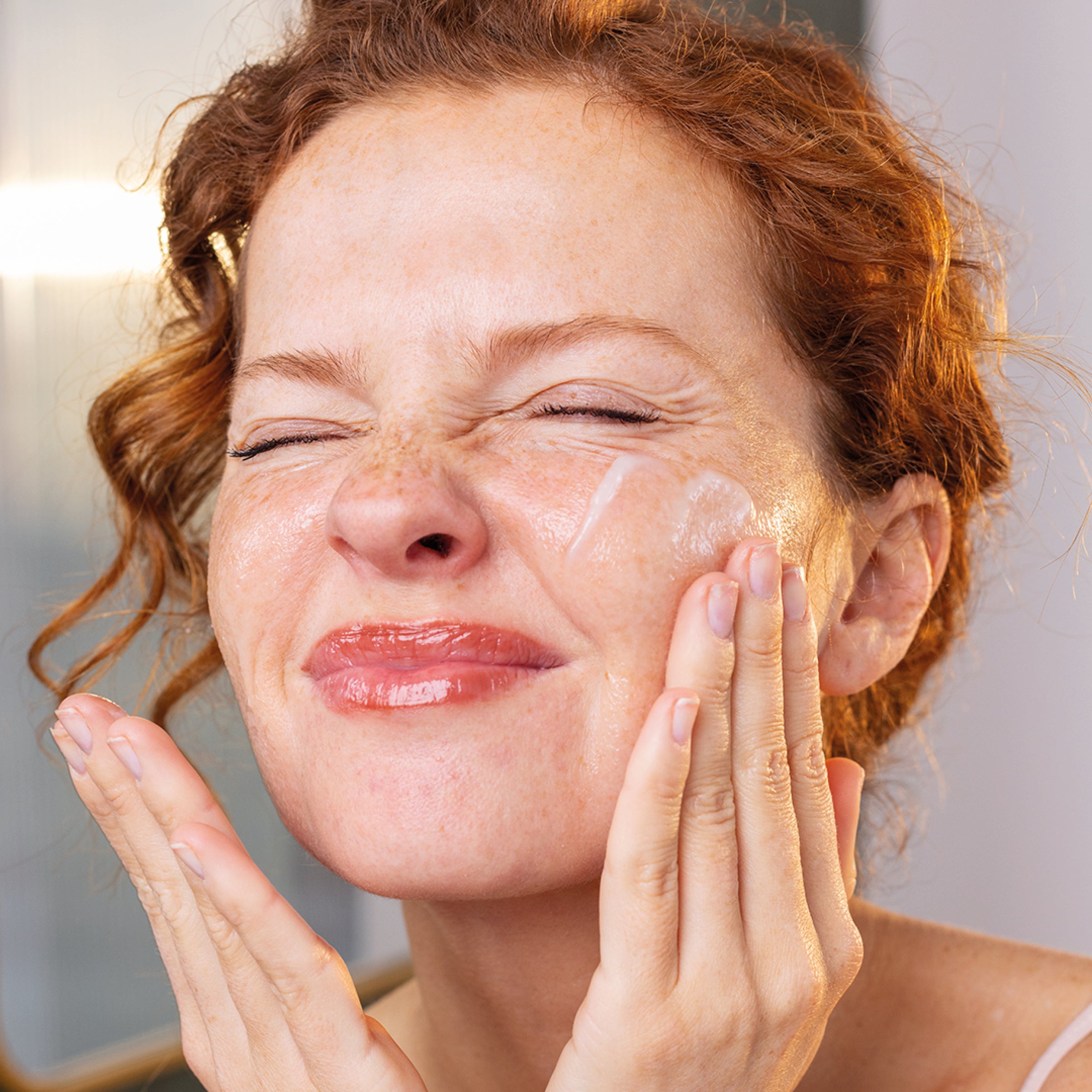 Woman with red hair applying SENSIFINE balm-to-oil cleanser to her face, showing a smooth, milky texture on her cheek.