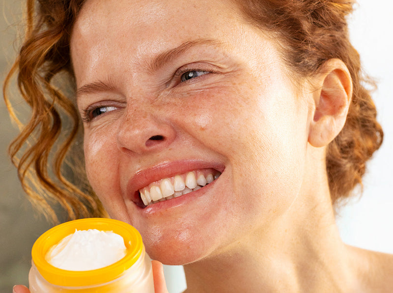 Smiling woman with curly red hair holding an open jar of white collagen booster cream.