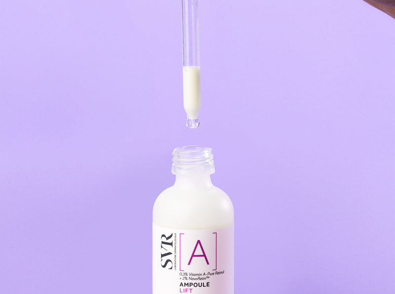 Hand holding a dropper over a bottle of SVR Vitamin A retinol serum against a soft purple background.