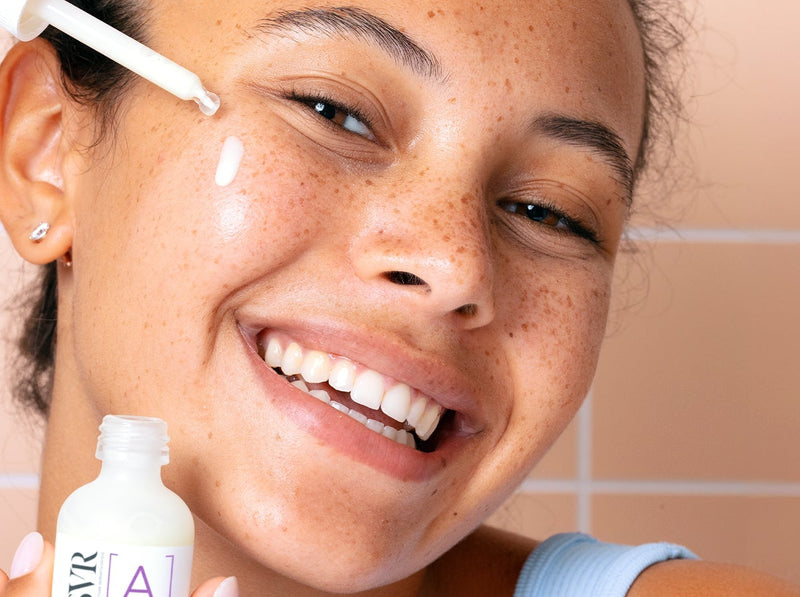 Smiling woman with freckles applying SVR retinol serum with a dropper to her cheek against a tiled background.