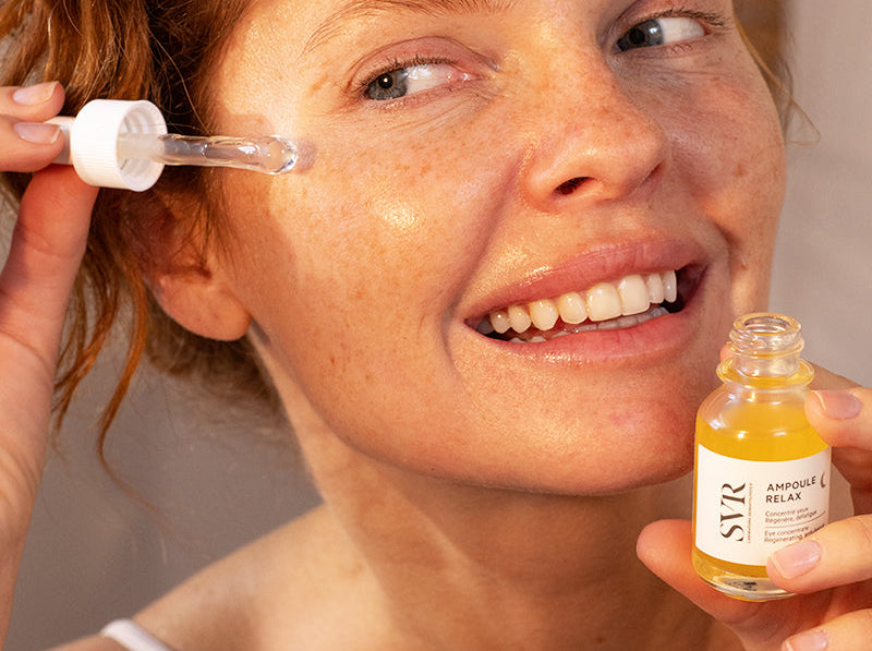 Smiling woman with freckles applying golden SVR eye serum with a dropper for healthy, glowing skin.