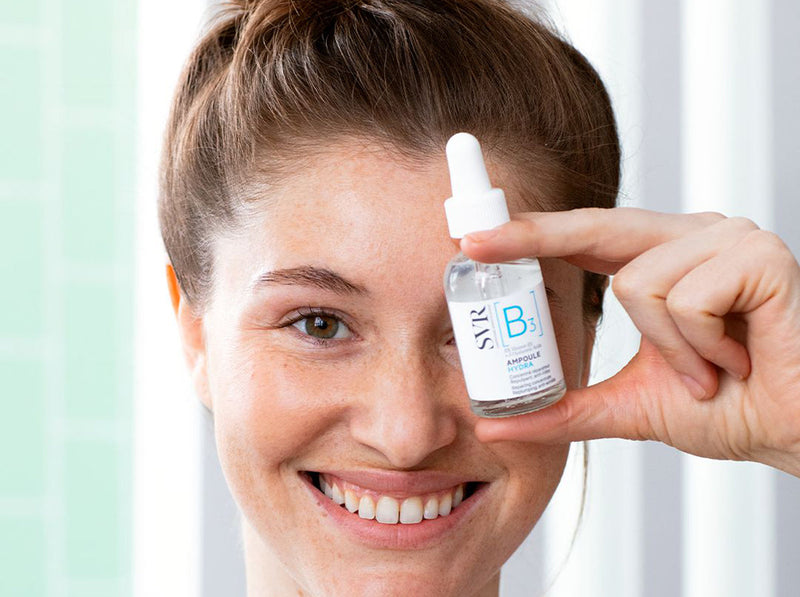 Smiling woman holding a clear SVR B3 hydrating serum bottle near her eye.