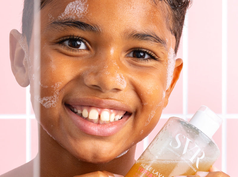 Smiling child with soap suds on face holding a bottle of SVR Topialyse cleansing oil in a pink tiled shower.