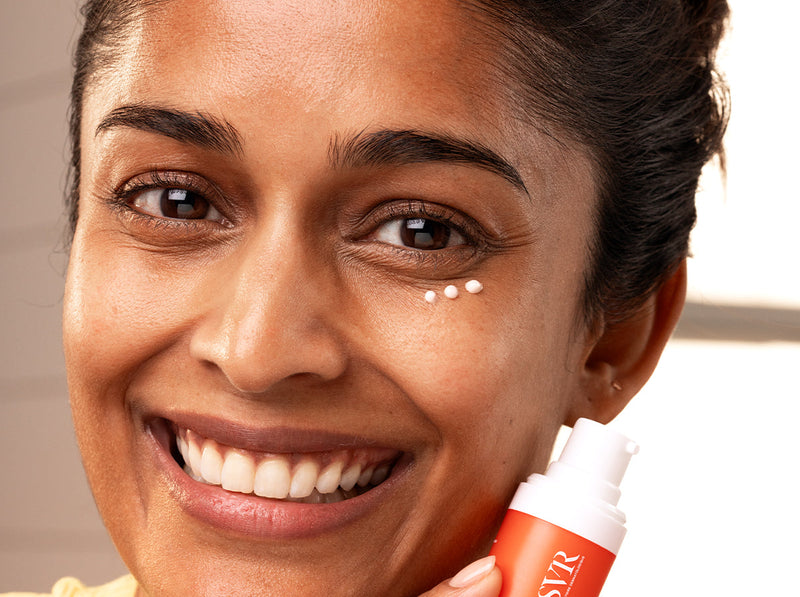 Smiling woman applying three small dots of white eye cream under her eye while holding an orange SVR bottle.