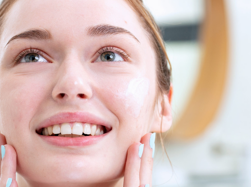 Young woman with clear skin applying a lightweight white moisturizer to her cheek in a bright bathroom.