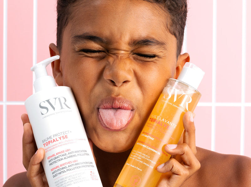 Young boy making a playful face while holding SVR skincare bottles for eczema relief against a pink tiled background.