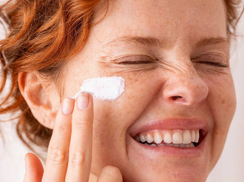 Smiling woman with red hair and freckles applying a white exfoliating face cream to her cheek.