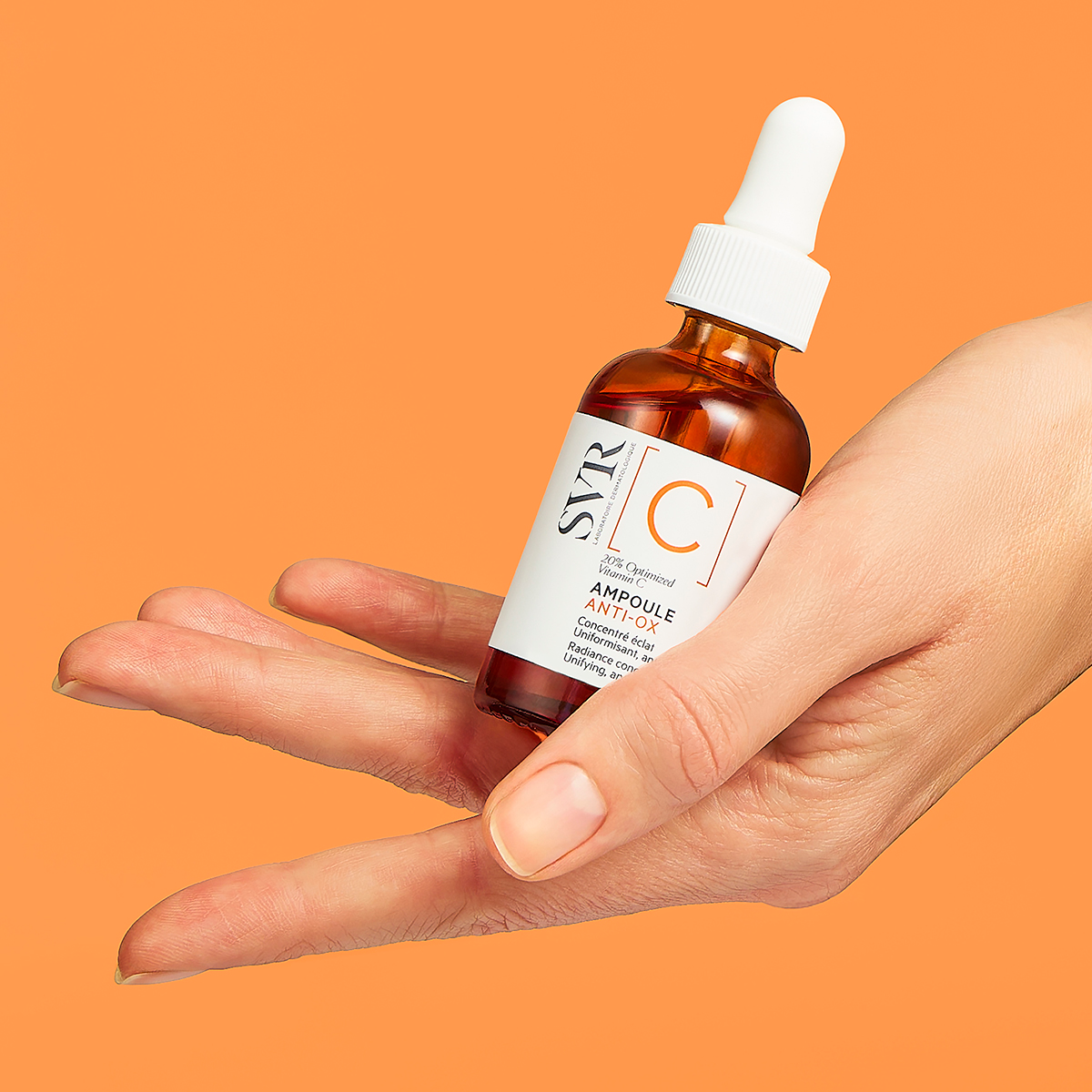 Hand holding an amber glass bottle of SVR Ampoule [C] Anti-Ox Vitamin C serum with a white dropper against an orange background.