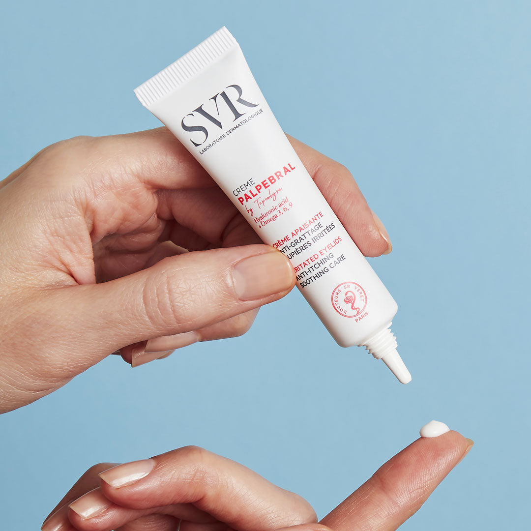 Hand holding a white SVR Palpebral eye cream tube, dispensing a small drop of white cream onto a fingertip.