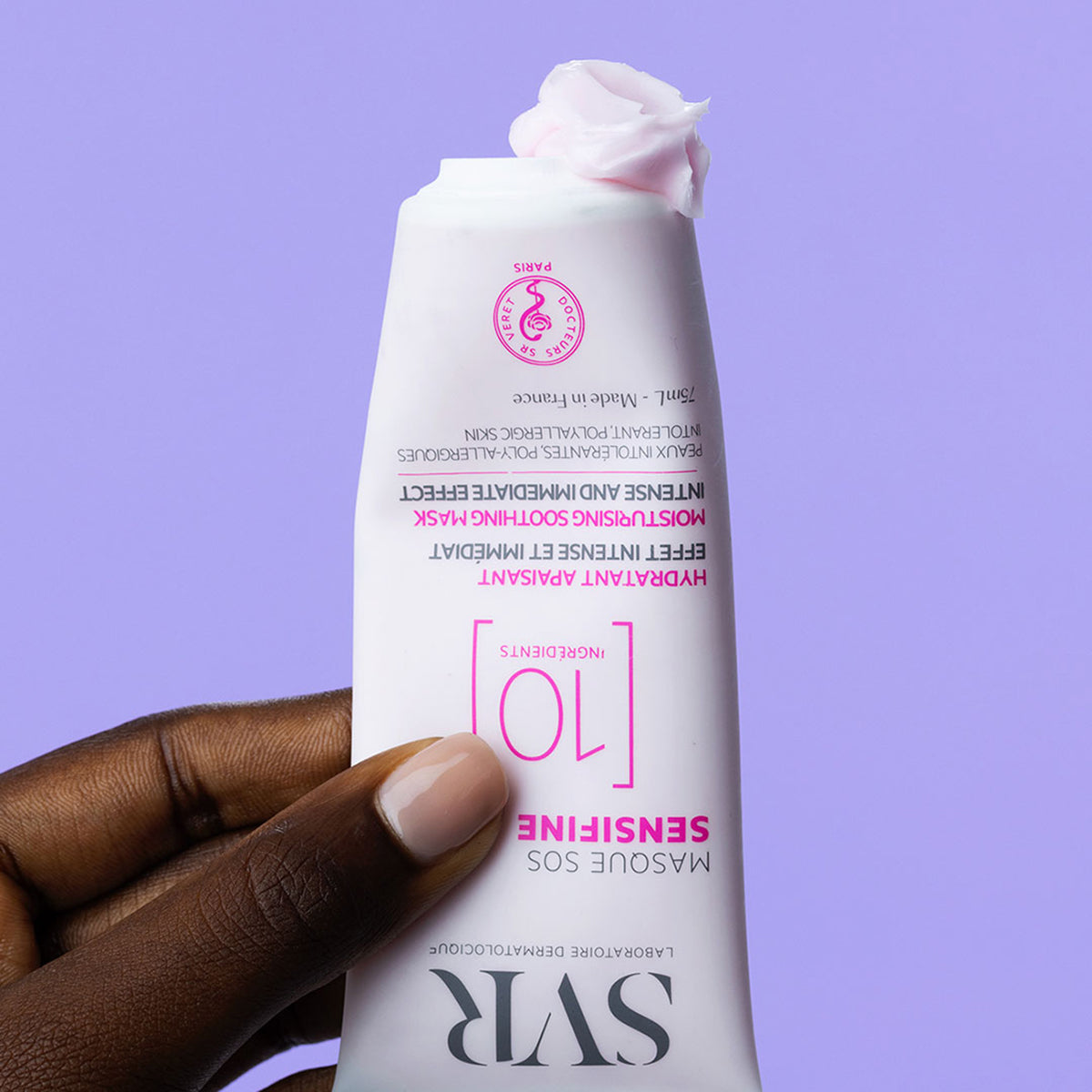 Hand holding a 75ml SVR Sensifine SOS mask tube with a dollop of pale pink cream against a light purple background.