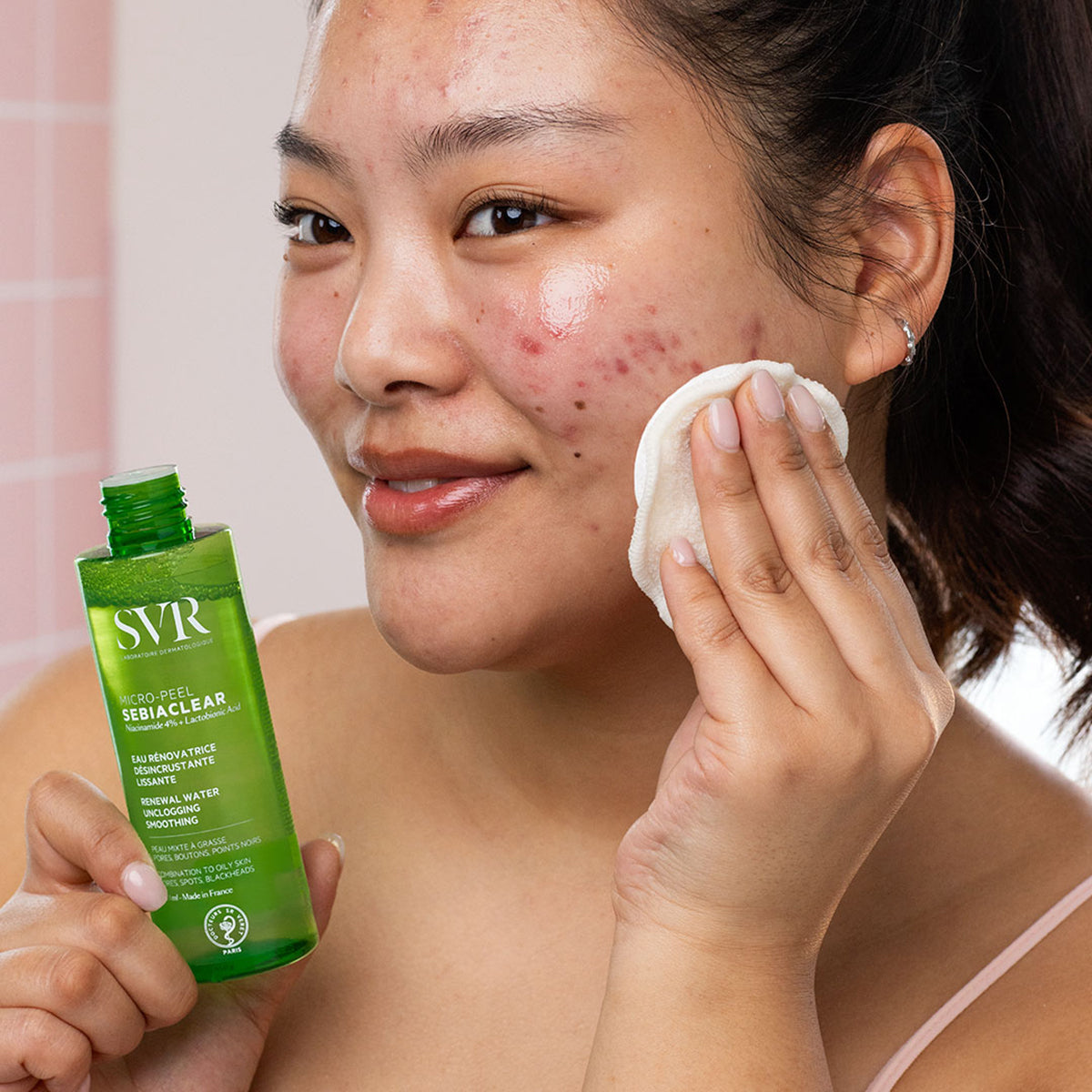 Woman applying SVR Sebiaclear Micro-Peel Essence with a cotton pad to her face to treat acne and skin texture.
