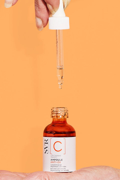 Hand holding a dropper over an amber glass bottle of SVR Ampoule Anti-Ox Vitamin C serum against an orange background.