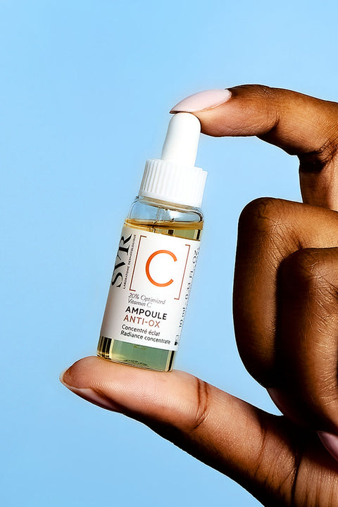 Hand holding a small SVR Ampoule Anti-Ox vitamin C serum bottle against a light blue background.