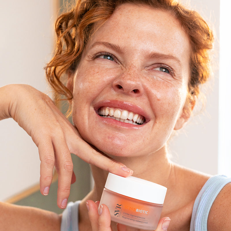 Femme rousse souriante tenant un pot de crème SVR Biotic C20 pour un teint éclatant et une peau lumineuse.