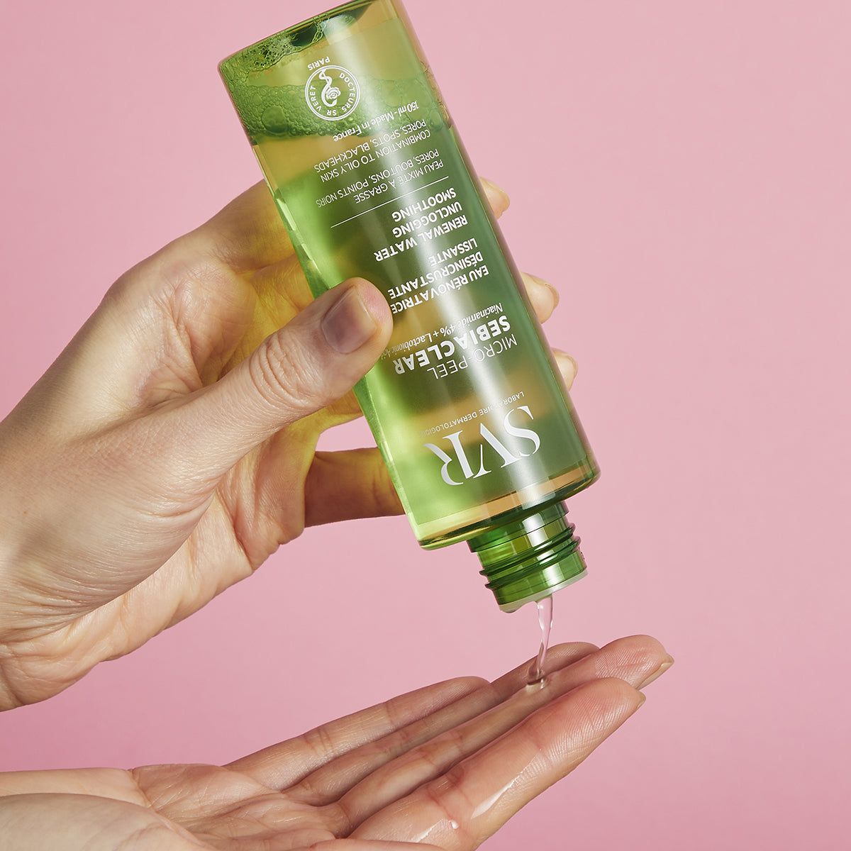 SVR Sebiaclear Micro-Peel green bottle pouring clear liquid into a hand against a soft pink background.