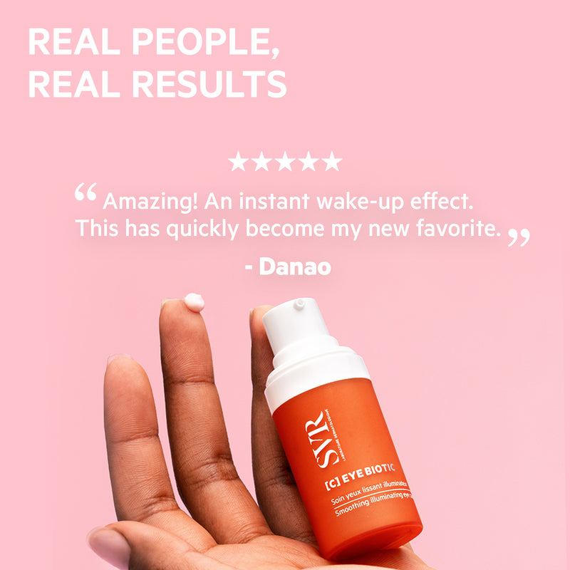 Hand holding an orange SVR [C] Eye Biotic pump bottle with a dab of cream on a fingertip against a pink background.