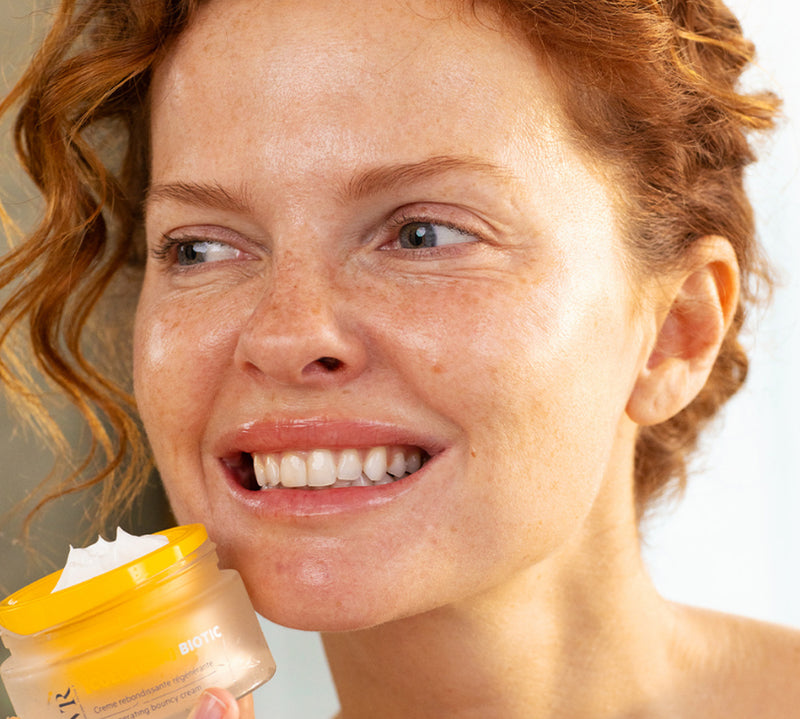 Open jar of yellow collagen face cream held by smiling red-haired woman with clear skin.