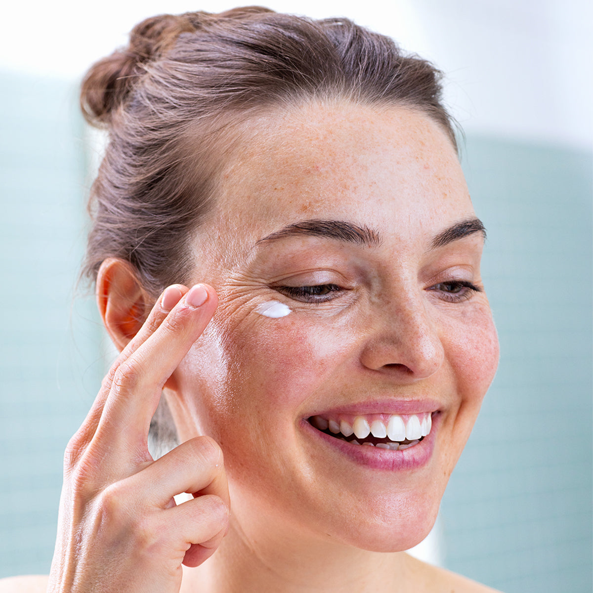 Smiling woman applying white SVR Palpebral eye cream to her sensitive under-eye area with her fingertip.