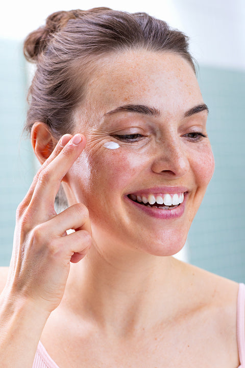 Smiling woman with freckles applying a small dab of white moisturizing eye cream to her under-eye area.