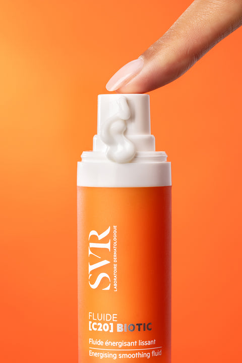 Finger pressing a white pump bottle of SVR [C20] Biotic energizing smoothing fluid against an orange background.