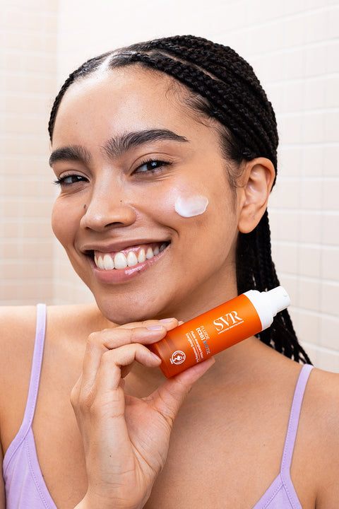 Smiling woman holding an orange SVR Fluide C20 bottle with a dab of cream on her cheek.