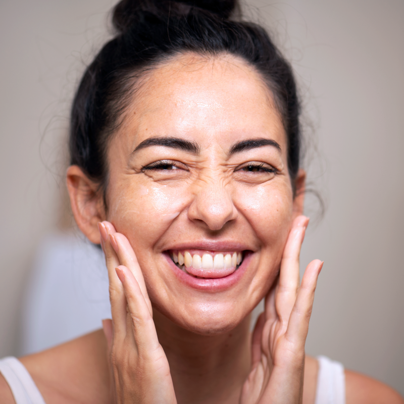 Smiling woman with glowing skin gently touching her cheeks, showcasing radiant complexion.