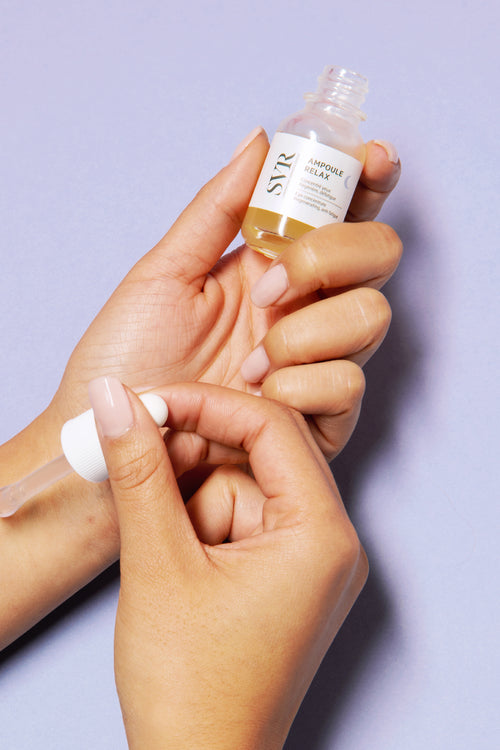 Hand holding open dropper bottle of Relax Eye Concentrate with serum drops on wrist against light purple background.