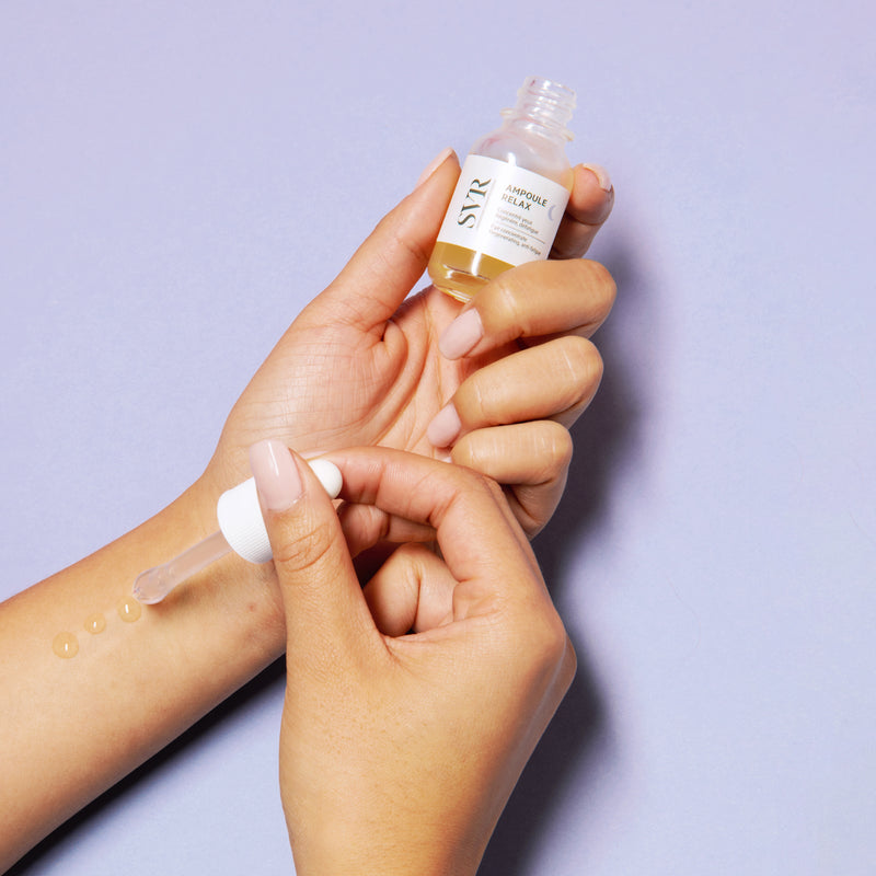 Hand holding open dropper bottle of Relax Eye Concentrate with serum drops on wrist against light purple background.