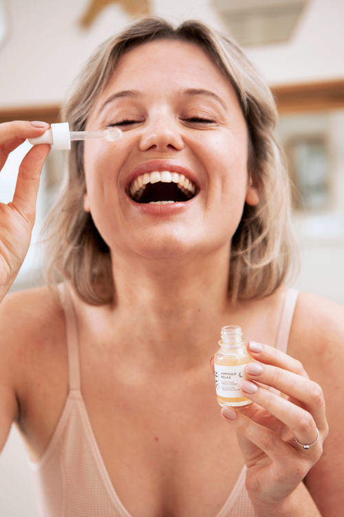 Woman holding a small dropper bottle of yellow-tinted eye serum, applying product near cheek with blurred background.