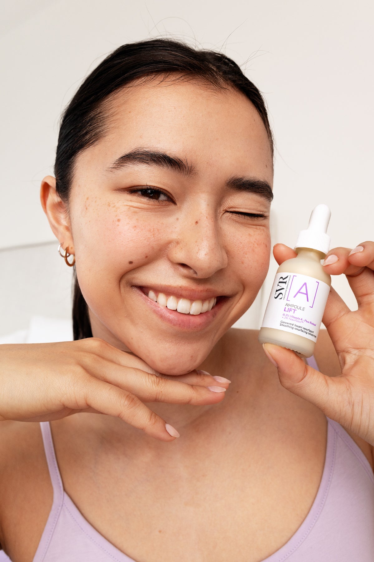 Smiling woman winking while holding a bottle of SVR Ampoule Lift retinol serum against a neutral background.