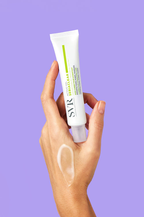 Hand holding a white tube of SVR Sebiaclear Active Gel with a translucent gel swatch on the skin against a purple background.